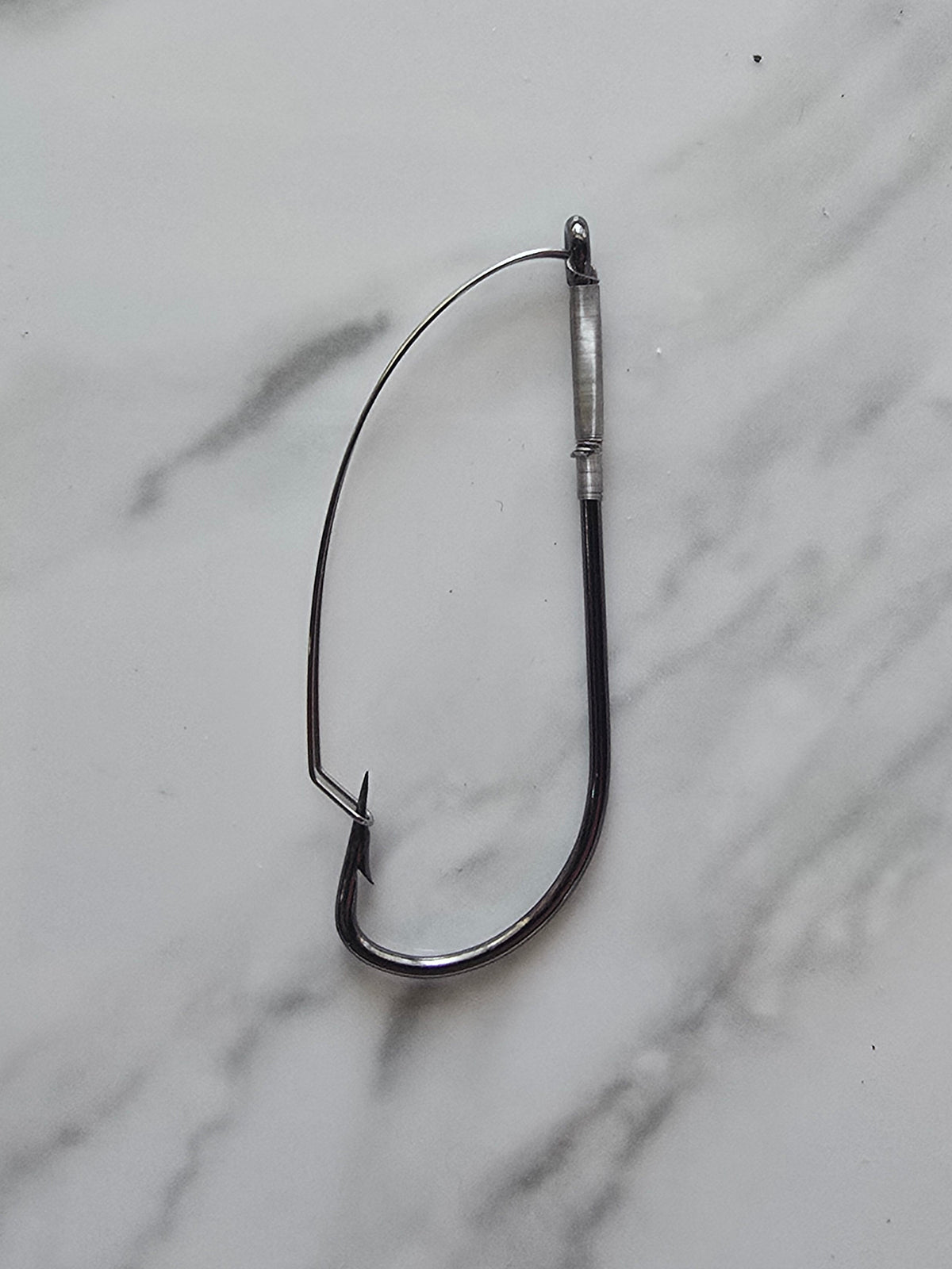 Royal Weed Guard Bait Hook A single black metal fishing hook with a silver metal eyelet and binding, placed on a white and gray marble surface.
